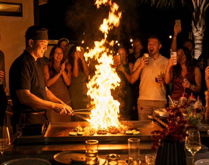 Hibachi chef creating a controlled fire show while guests cheer at night