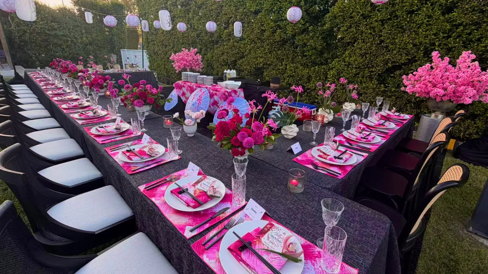 Outdoor birthday dinner table with pink floral decorations, hanging lanterns, and place settings in a garden.